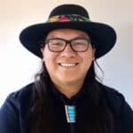 Headshot of a man with long dark hair, glasses, a brimmed hat, looking at the camera and smiling