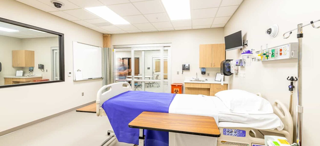 Simulation nursing exam room with hands-on learning equipment and a one-way mirror.